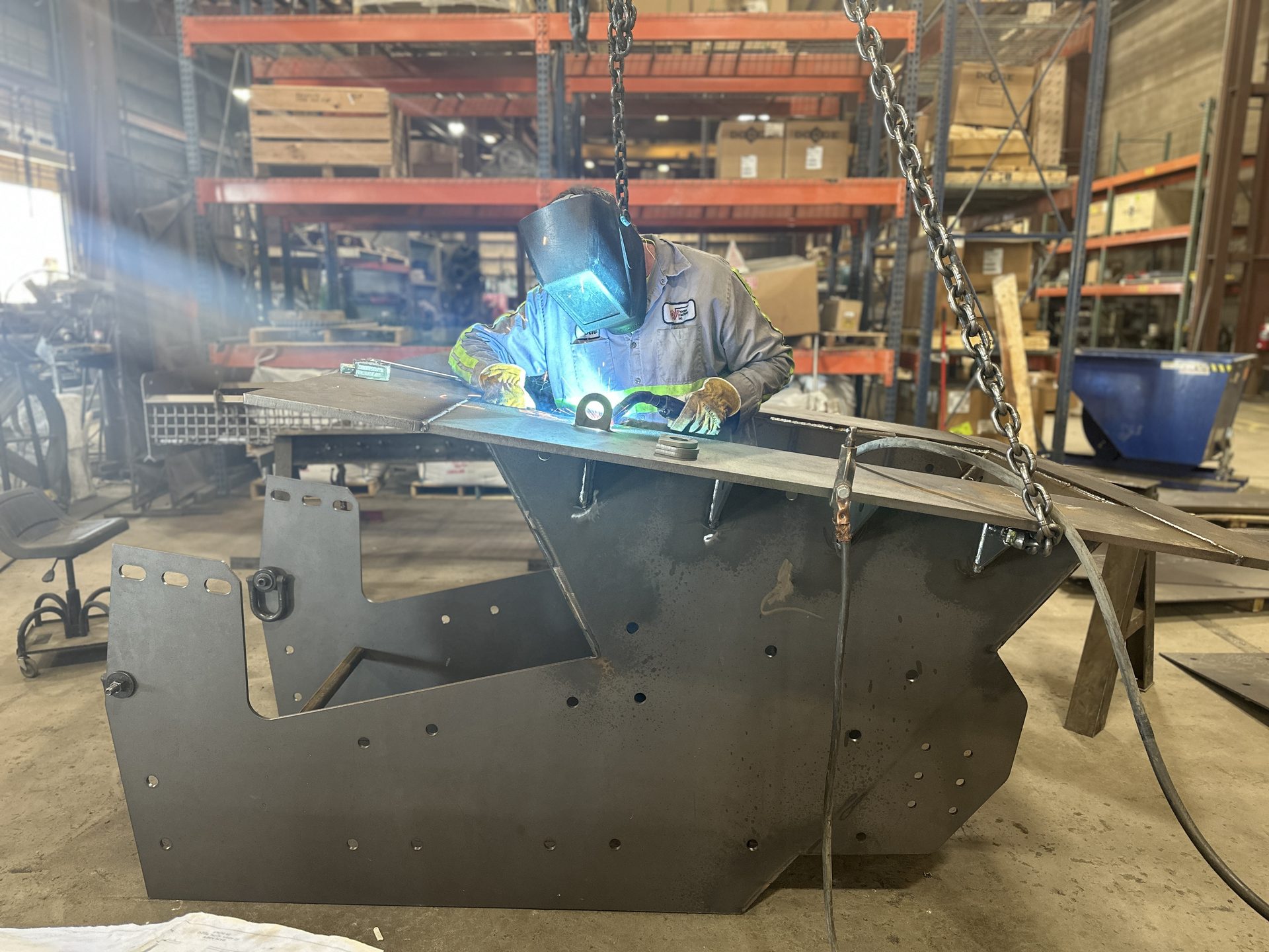 Welder in a welding helmet welding a large cut-steel chute in the PMI shop
