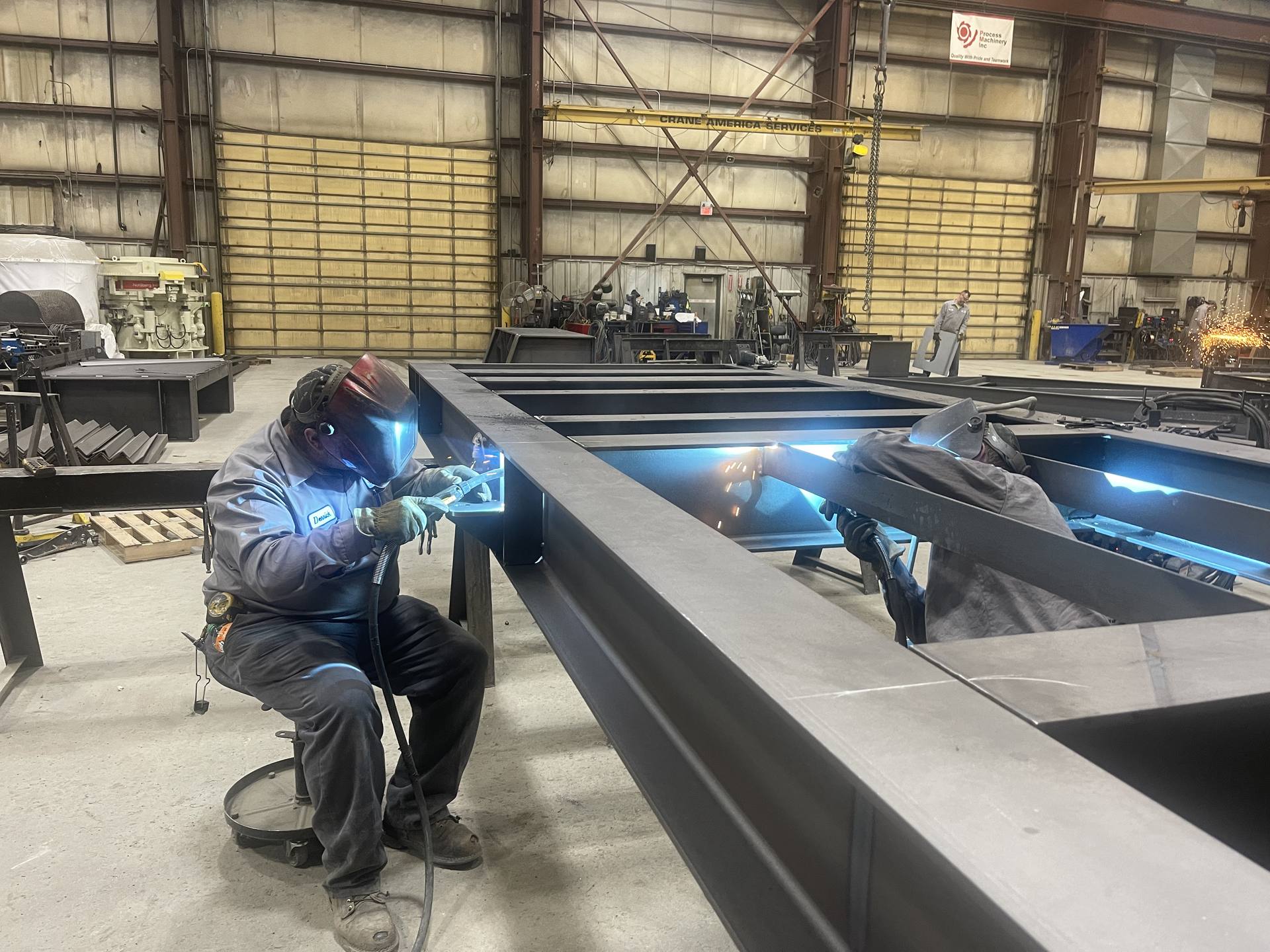 Two welders in helmets welding a large steel frame on the PMI shop floor